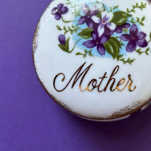 Op de afbeelding: Witte keramische sieradenbox met een gouden rand en een bloemmotief met paarse en witte bloemen. De doos heeft het woord "Mother" in gouden letters.