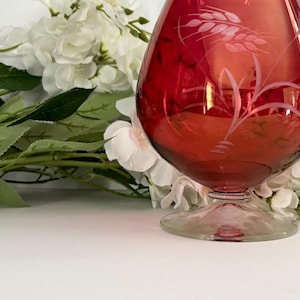 May include: A red glass vase with an etched wheat design. The vase has a round body and a flared lip.
