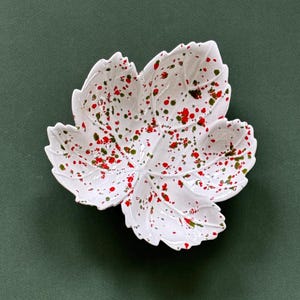 Op de afbeelding: Witte keramische blad-vormige schaal met rode en groene spikkels. De schaal heeft een gedetailleerd bladnerf ontwerp en staat tegen een donkergroene achtergrond. Ideaal voor het serveren van snacks of als decoratief element.
