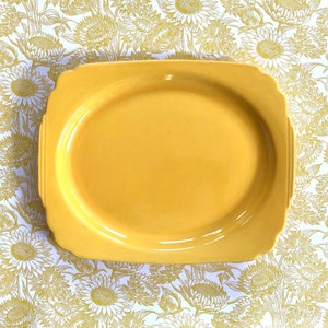 May include: A yellow ceramic serving platter with a slightly raised rim and a scalloped edge. The platter is on a white background with a floral pattern.