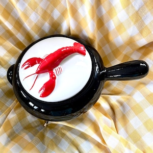 May include: A black ceramic serving dish with a white interior featuring a vibrant red lobster design. The dish has a long handle and a small foot. The background is a yellow and white checkered pattern.