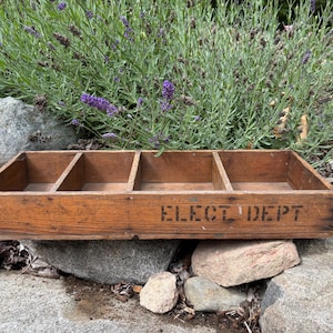 Puede incluir: Una caja de madera vintage con cuatro compartimentos, con el texto estampado "ELECT DEPT" en negro. La caja es de madera envejecida, mostrando signos de desgaste. La caja descansa sobre una superficie de piedra, con plantas de lavanda en el fondo.