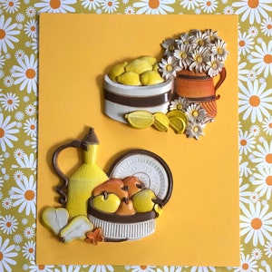 Könnte beinhalten: Ein gelbes und braunes Wanddekor-Set mit einem Krug, einer Obstschale und einer Vase mit Blumen. Die Obstschale ist mit Zitronen gefüllt und die Vase mit weißen Gänseblümchen.