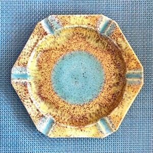 May include: A hexagonal ashtray with a speckled design. The centre is a light blue circle, surrounded by a brown and yellow speckled pattern. The rim has four light blue indentations for resting cigarettes.