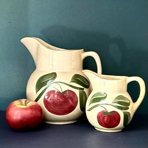 Puede incluir: Dos jarras de cerámica color crema con diseños de manzanas rojas y hojas verdes. Una manzana roja está delante de las jarras. Las jarras tienen asas y picos. El fondo es de color verde azulado.