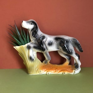 May include: A ceramic planter shaped like a black and white dog with a green plant inside. The dog is standing on a yellow and orange base.
