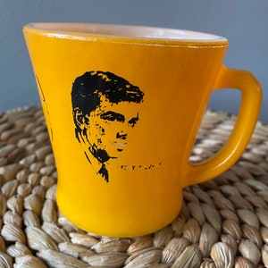 May include: Yellow ceramic mug with a black portrait of a man and the text "E. Y. S.". The mug has a curved shape and a yellow handle. The interior of the mug is white.