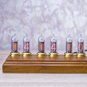Op de afbeelding: Een houten klok met zes heldere glazen Nixie-buizen die de tijd "14:14:10" weergeven. De buizen hebben oranje cijfers op een donkere achtergrond. De klok staat op een licht houten oppervlak.
