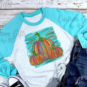 Puede incluir: Una camisa de manga larga blanca y turquesa con un dise&ntilde;o de calabaza colorido. La camisa tiene una manga turquesa y una manga blanca con un dise&ntilde;o de calabaza en la manga blanca. El dise&ntilde;o de la calabaza es un dise&ntilde;o abstracto colorido con colores naranja, amarillo y verde.