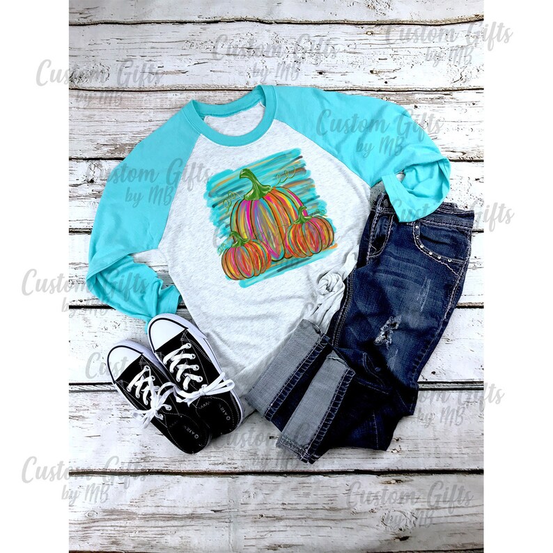 Puede incluir: Una camisa de manga larga blanca y turquesa con un dise&ntilde;o de calabaza colorido. La camisa tiene una manga turquesa y una manga blanca con un dise&ntilde;o de calabaza en la manga blanca. El dise&ntilde;o de la calabaza es un dise&ntilde;o abstracto colorido con colores naranja, amarillo y verde.
