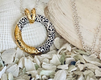 May include: A silver and gold pendant necklace with a circular design. The pendant has a textured, hammered gold finish on one side and a silver filigree design on the other side. The necklace is on a silver chain.