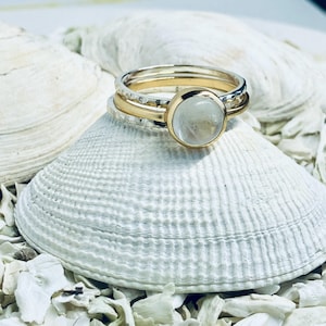 May include: Three rings, two silver and one gold, are stacked together. The top ring has a textured design, while the middle ring is gold. The top ring features a large, round, clear gemstone set in a gold bezel. The rings are displayed on a white seashell.