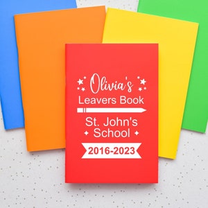 May include: A stack of notebooks in red, orange, yellow, green and blue. The top notebook is red and has the text "Olivia's Leavers Book St. John's School 2016-2023" printed on it.