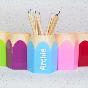 May include: Five colourful pencil-shaped desk organisers. The organisers are in green, purple, blue, pink, and red. The blue organiser has the name "Archie" in white. Pencils are placed in the blue organiser.
