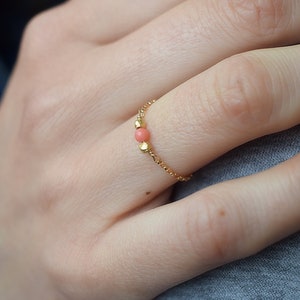 Puede incluir: Un delicado anillo de cadena de oro con una sola cuenta de coral rosa. El anillo se lleva en un dedo.