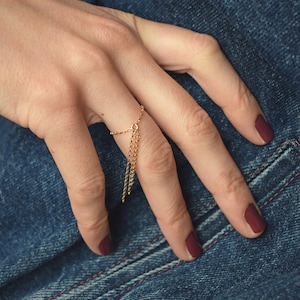 Könnte beinhalten: Ein goldener Kettenring mit einem baumelnden Kettendetail. Der Ring wird an einem Finger mit burgunderfarbenem Nagellack getragen.