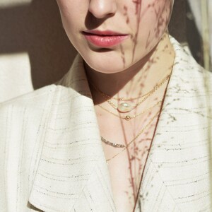 Op de afbeelding: Een vrouw draagt een wit blazer met een gelaagde gouden ketting met een parel hanger. De ketting is gestyled met een delicate gouden ketting en een kleine gouden kraal.