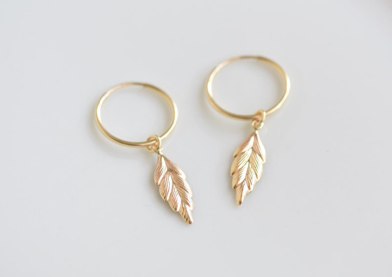 May include: A pair of gold hoop earrings with a small gold leaf charm dangling from each hoop.