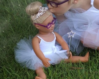 May include: A doll dressed as a ballerina, wearing a white leotard and tutu, purple glasses, and a tiara. The doll is holding a silver star wand. Another person is kissing the doll on the head. The scene is set on green grass.