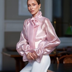 Könnte beinhalten: Eine hellrosa, transparente Bluse mit hohem Kragen und großer Schleife. Die langen Ärmel haben gerüschte Manschetten. Die Bluse wird mit einer weißen Hose kombiniert. Der Gesamtstil ist elegant und anspruchsvoll.