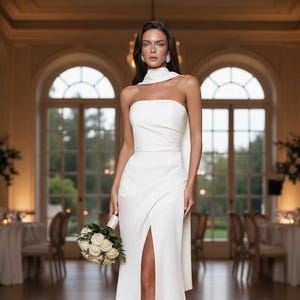 Puede incluir: Vestido de novia blanco sin tirantes con una abertura alta, un escote drapeado y una falda fluida. La novia sostiene un ramo de rosas blancas. El vestido se usa en una habitación bien iluminada con grandes ventanas arqueadas.