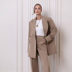 Puede incluir: Un traje pantalón beige con una blazer larga de dos botones y pantalones de pernera ancha a juego. El traje se combina con una camiseta blanca y un collar con colgante dorado. El conjunto es de color neutro y tiene un corte a medida.