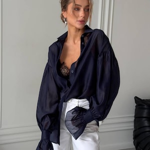 May include: A dark navy blue blouse with long, voluminous sleeves and ruffled cuffs. The blouse is partially unbuttoned, revealing a black lace undergarment. The model is wearing white pants. The blouse has a classic collar.