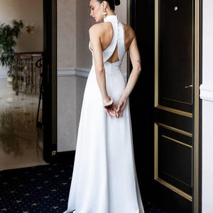 K&ouml;nnte beinhalten: Ein wei&szlig;es Brautkleid mit Neckholder-Ausschnitt, offenem R&uuml;cken und flie&szlig;endem Rock. Das Kleid hat einen hohen Ausschnitt und ein schlichtes, minimalistisches Design. Die Frau tr&auml;gt Ohrringe und ein Armband. Der Hintergrund umfasst eine dunkle T&uuml;r mit goldenen Zierleisten und einen gemusterten Teppich.