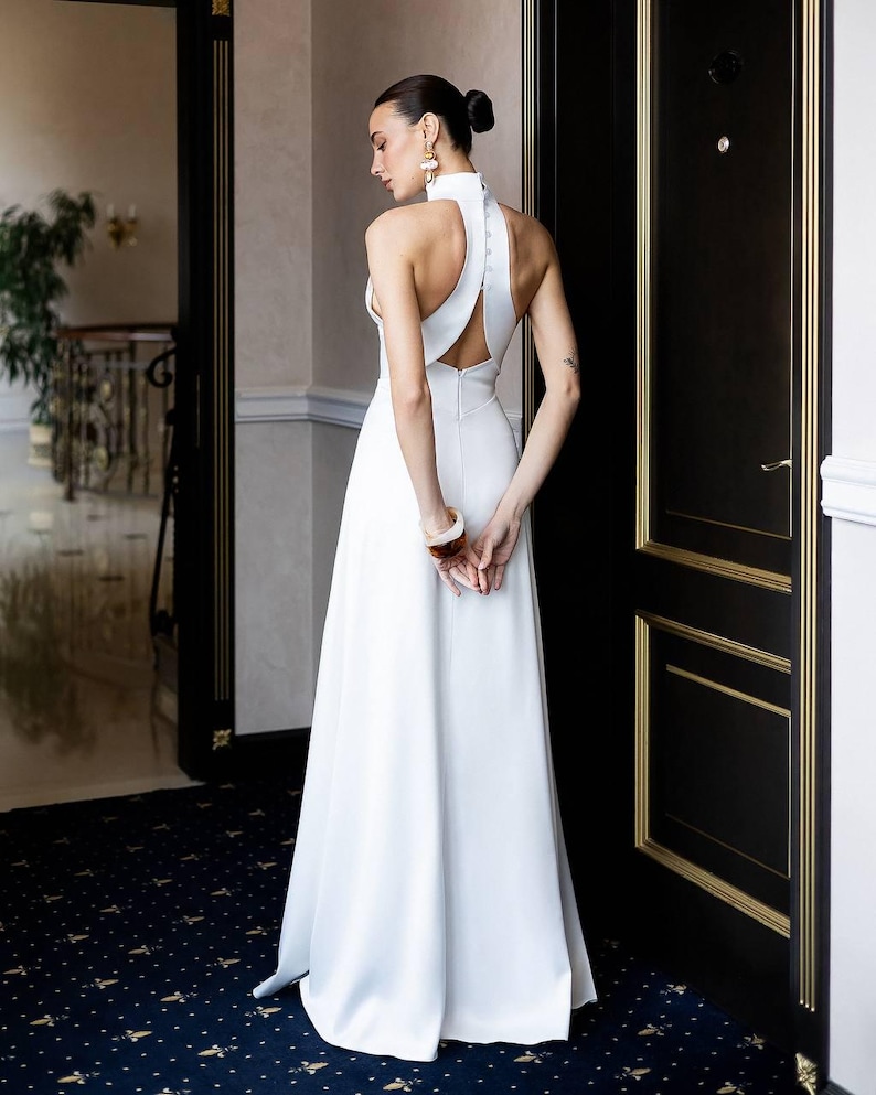 K&ouml;nnte beinhalten: Ein wei&szlig;es Brautkleid mit Neckholder-Ausschnitt, offenem R&uuml;cken und flie&szlig;endem Rock. Das Kleid hat einen hohen Ausschnitt und ein schlichtes, minimalistisches Design. Die Frau tr&auml;gt Ohrringe und ein Armband. Der Hintergrund umfasst eine dunkle T&uuml;r mit goldenen Zierleisten und einen gemusterten Teppich.