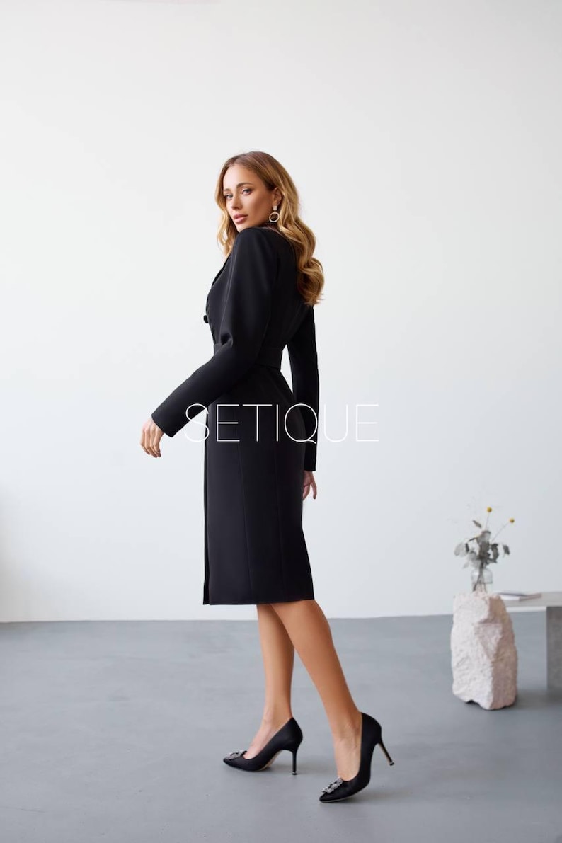 Peut inclure: Une robe noire avec un haut de style blazer et une jupe mi-longue. La robe a une taille ceintur&eacute;e et des boutons. L'image comprend le texte "SETIQUE". Le mod&egrave;le porte des talons hauts noirs.