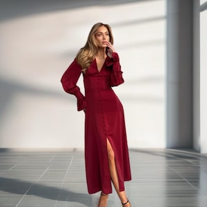 May include: A long, burgundy dress with long sleeves and a V-neck. The dress has a button-down front and a high slit on the left side. The model is wearing black strappy heels.