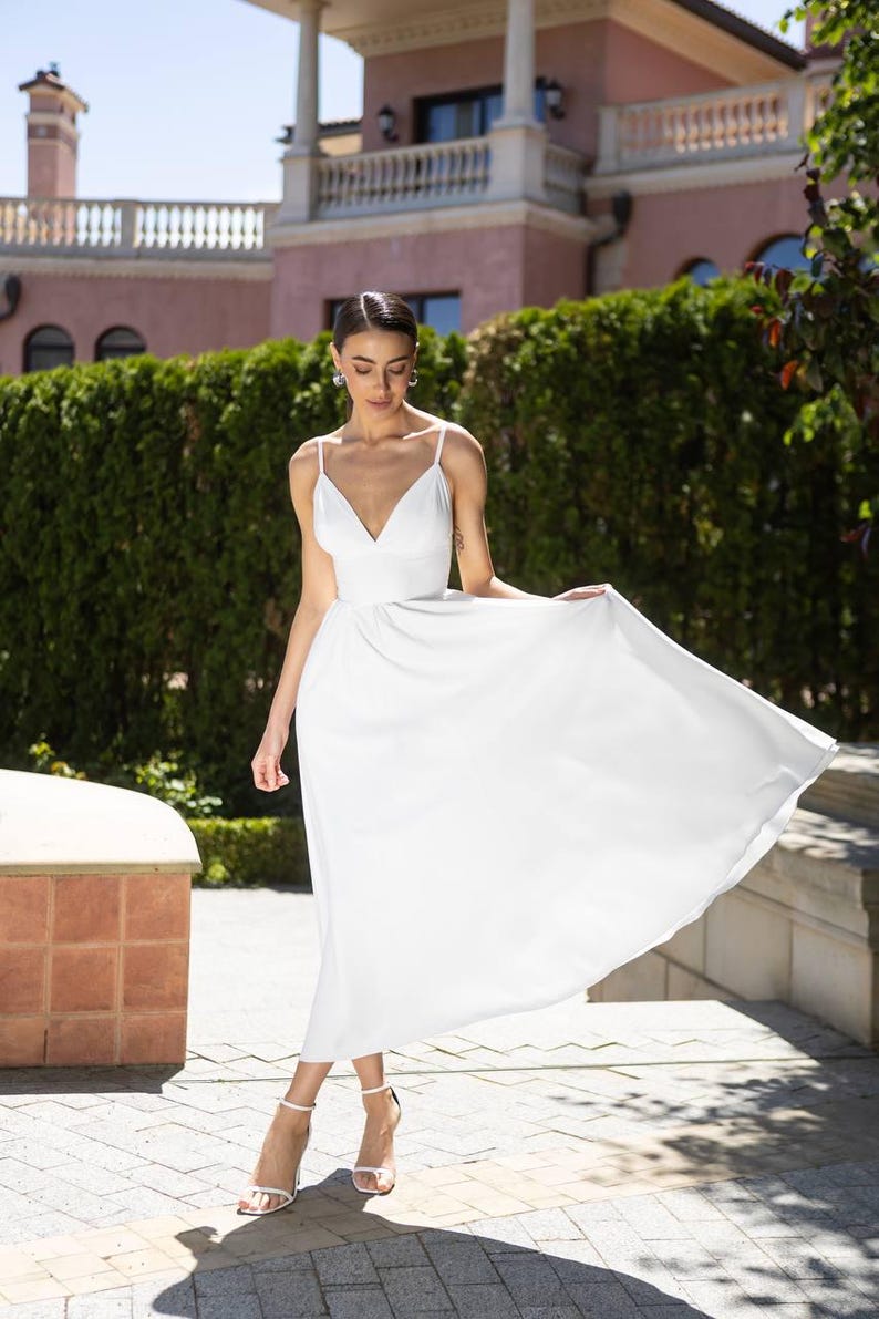 May include: A white, flowing dress with spaghetti straps and a V-neck. The dress is knee-length and appears to be made of a lightweight fabric. The model is wearing white heels. The background includes a green hedge and a pink building.