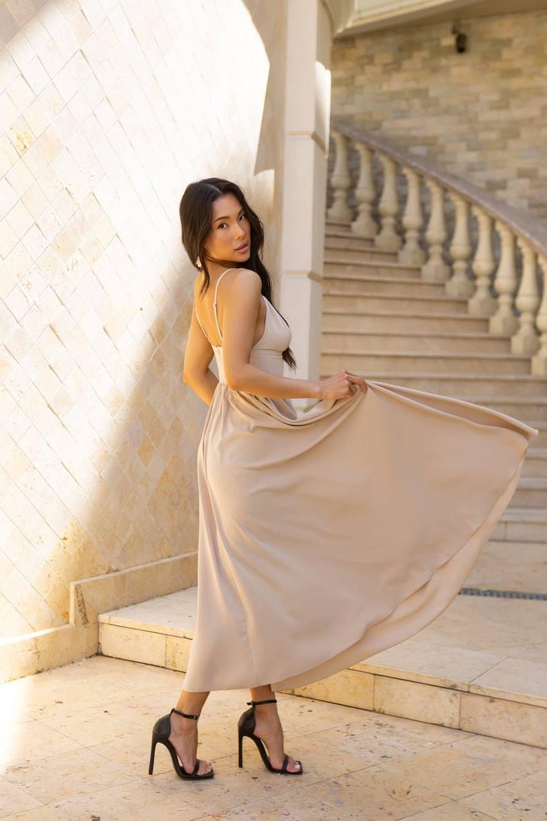 May include: A beige maxi dress with spaghetti straps and a flowing skirt. The dress is paired with black high-heeled sandals. The background features a stone wall and a staircase with a decorative railing.