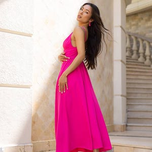 May include: A vibrant fuchsia-colored maxi dress with a flowing silhouette. The dress features a spaghetti strap neckline and is paired with black high-heeled sandals. The model is wearing pink earrings. The background includes a white wall and a staircase.