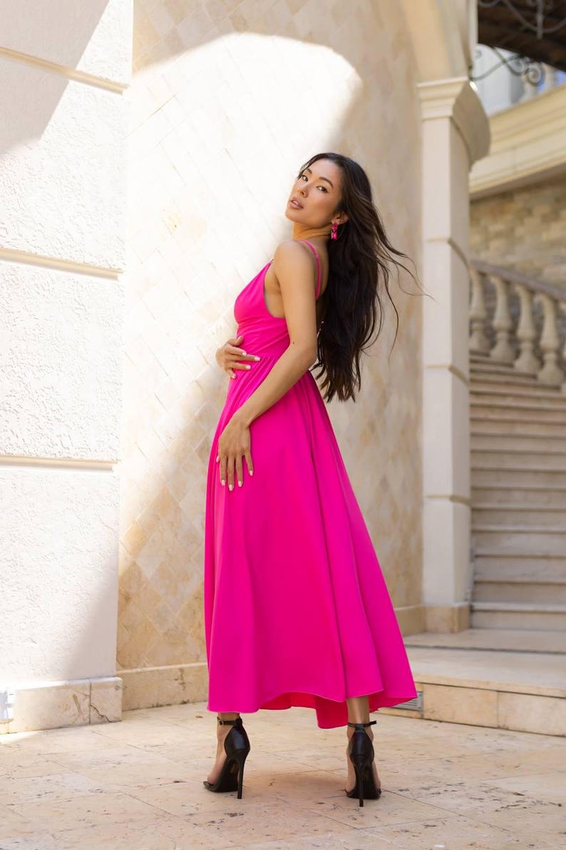 May include: A vibrant fuchsia-colored maxi dress with a flowing silhouette. The dress features a spaghetti strap neckline and is paired with black high-heeled sandals. The model is wearing pink earrings. The background includes a white wall and a staircase.