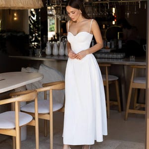Puede incluir: Un vestido blanco largo hasta el suelo con un escote coraz&oacute;n estructurado, tirantes finos y un corpi&ntilde;o ajustado que se transforma en una falda amplia y fluida. La prenda se combina con zapatos de tac&oacute;n blancos con tira trasera.