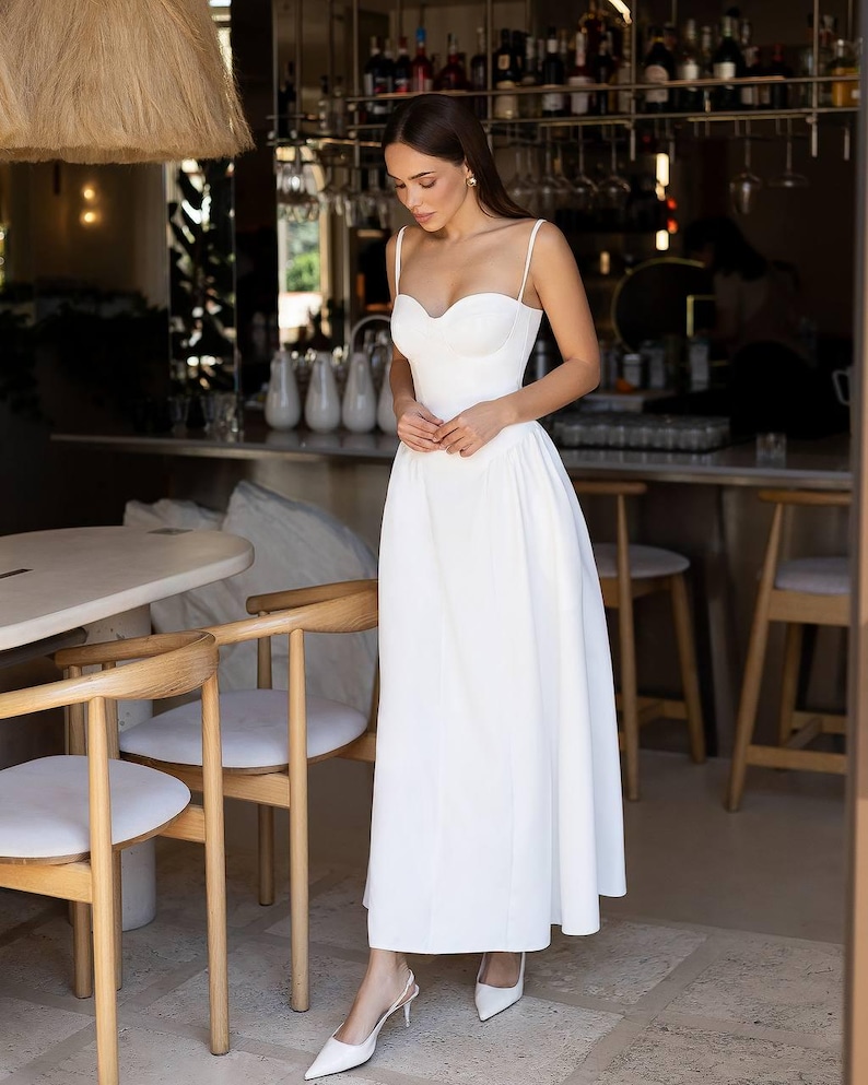Puede incluir: Un vestido blanco largo hasta el suelo con un escote coraz&oacute;n estructurado, tirantes finos y un corpi&ntilde;o ajustado que se transforma en una falda amplia y fluida. La prenda se combina con zapatos de tac&oacute;n blancos con tira trasera.