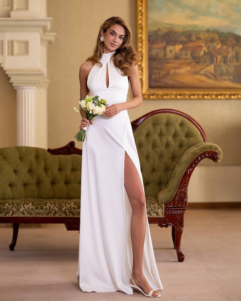 K&ouml;nnte beinhalten: Ein wei&szlig;es Neckholder-Kleid mit hohem Schlitz. Das Kleid hat einen Schl&uuml;sselloch-Ausschnitt und wird mit wei&szlig;en Sandalen getragen. Die Person h&auml;lt einen Strau&szlig; wei&szlig;er und gr&uuml;ner Blumen. Im Hintergrund befinden sich ein gr&uuml;nes Sofa und ein Gem&auml;lde.
