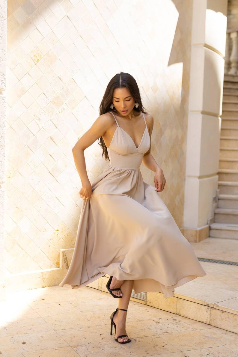 May include: A beige spaghetti strap dress with a V-neck and a flowing skirt. The dress is paired with black high-heeled sandals. The woman is standing in front of a stone wall and stairs.