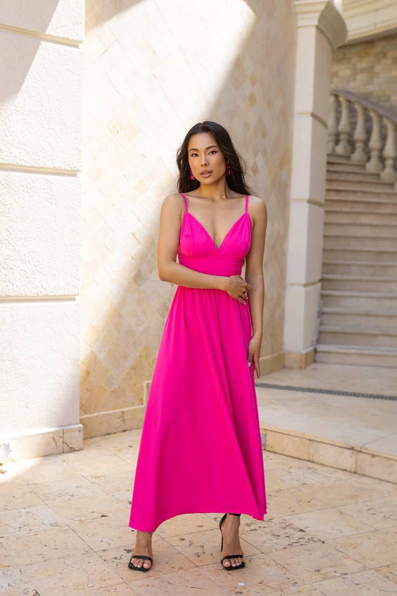 May include: A vibrant fuchsia maxi dress with a V-neckline and spaghetti straps. The dress has a flowing, A-line skirt and is paired with black heeled sandals. The background features a stone wall and staircase.