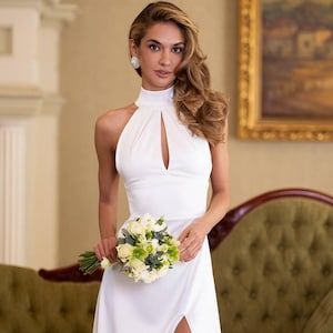 K&ouml;nnte beinhalten: Wei&szlig;es Brautkleid mit Halfter-Ausschnitt, Schl&uuml;sselloch-Ausschnitt und hohem Schlitz. Die Braut h&auml;lt einen Strau&szlig; wei&szlig;er und gr&uuml;ner Blumen. Das Kleid ist bodenlang und hat ein schlichtes, elegantes Design.