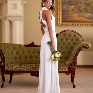 K&ouml;nnte beinhalten: Ein langes, wei&szlig;es Kleid mit Halfter-Ausschnitt und offenem R&uuml;cken. Die Person h&auml;lt einen Strau&szlig; wei&szlig;er Blumen. Der Hintergrund zeigt ein gr&uuml;nes Sofa und ein Gem&auml;lde in einem goldenen Rahmen. Das Kleid ist bodenlang.