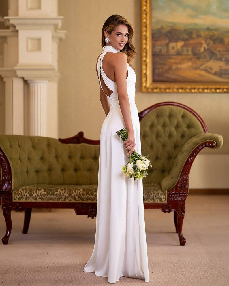 K&ouml;nnte beinhalten: Ein langes, wei&szlig;es Kleid mit Halfter-Ausschnitt und offenem R&uuml;cken. Die Person h&auml;lt einen Strau&szlig; wei&szlig;er Blumen. Der Hintergrund zeigt ein gr&uuml;nes Sofa und ein Gem&auml;lde in einem goldenen Rahmen. Das Kleid ist bodenlang.