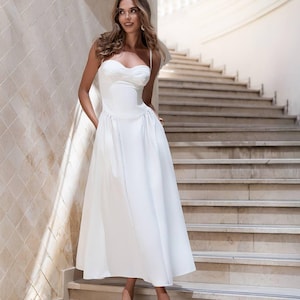 Puede incluir: Un vestido midi blanco que presenta un corpi&ntilde;o estructurado con escote coraz&oacute;n, tirantes finos y una falda amplia plisada. La prenda es de color blanco s&oacute;lido, confeccionada con un acabado liso e incluye una cintura ajustada que se transforma en una silueta acampanada.