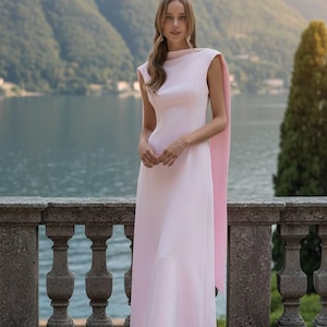 May include: A light pink, floor-length gown with a boat neckline and short sleeves. The dress features a flowing cape in a slightly darker shade of pink. The background includes a lake and mountains.