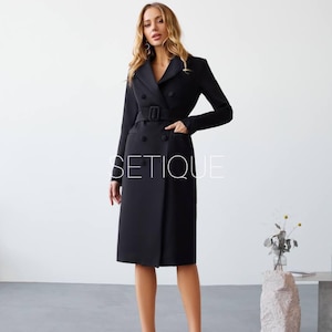 Peut inclure: Une robe manteau noire &agrave; double boutonnage avec une ceinture, des poches et des manches longues. La robe arrive au genou et a une coupe ajust&eacute;e. Le mot "SETIQUE" est imprim&eacute; sur la robe. Le mod&egrave;le porte des talons hauts noirs.