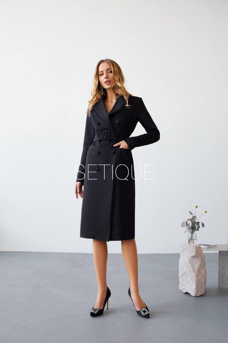 Peut inclure: Une robe manteau noire &agrave; double boutonnage avec une ceinture, des poches et des manches longues. La robe arrive au genou et a une coupe ajust&eacute;e. Le mot "SETIQUE" est imprim&eacute; sur la robe. Le mod&egrave;le porte des talons hauts noirs.