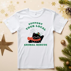 Może przedstawiać: Biała koszulka z grafiką czarnego kota w czerwonym szaliku i wieńcem z ostrokrzewu. Tekst brzmi "Support Your Local Animal Rescue" w kolorze zielonym. Projekt jest świąteczny i tematyczny.