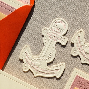 May include: An anchor-shaped invitation card in cream-colored cardstock with embossed details and red text. The text reads: ALL ABOARD THE PRE-KNOT YACHT, Friday March 27th, 11am-2pm, Join us for a day on the water onboard our private charter, Venue: Marina Cabo San Lucas | Attire: Swimwear.