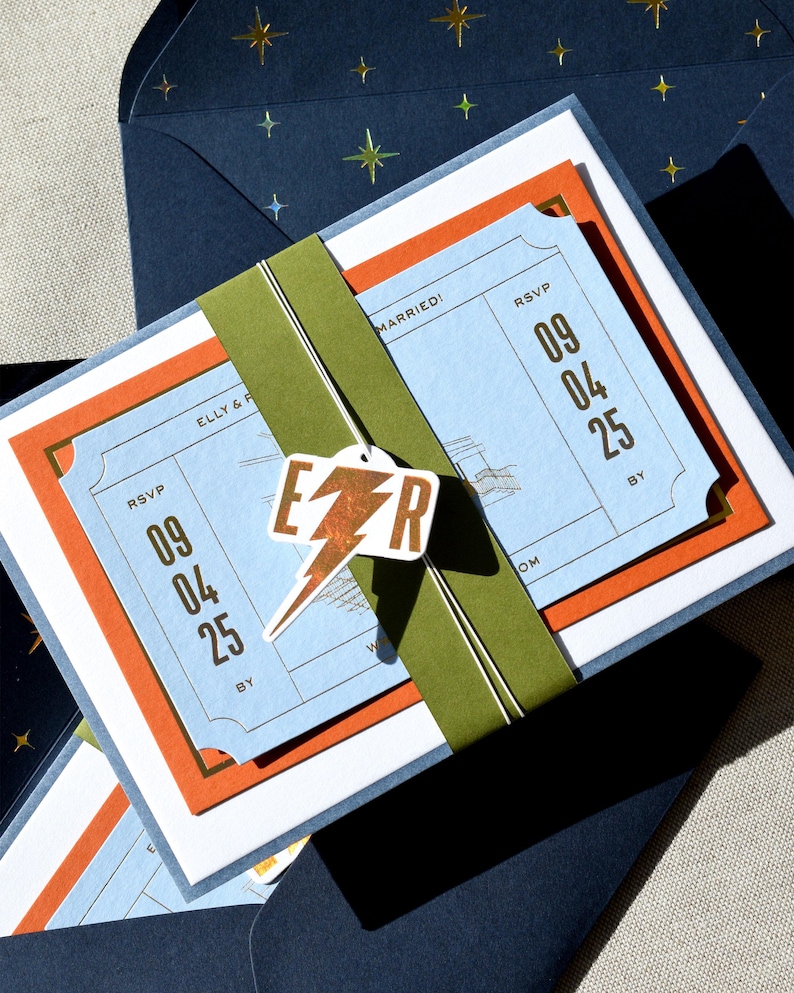 May include: A wedding invitation set featuring a light blue ticket-style card with the text ELLY & R and RSVP 09 04 25, layered over an orange card. An olive green paper band holds the set together, accented by a white tag with a copper lightning bolt and the letters E and R. The set is tucked into a dark blue envelope with a gold star pattern.
